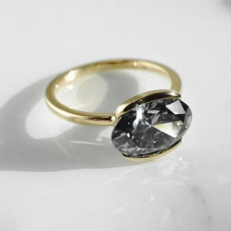 This Grey Moissanite Oval Cut Half Bezel Set Engagement Ring redefines contemporary style with clean lines and bold character. At its center sits a 3 carat dark grey moissanite oval cut gem, set in a sleek half bezel that runs east-west along the finger.